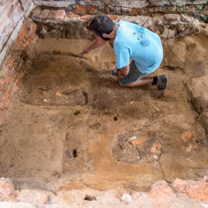 Kneeling archaeologist excavates dirt floor in front of brick wall