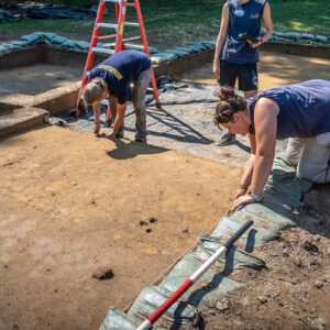 Archaeological Field Technicians Gabriel Brown and Hannah Barch prepare one of the squares at the clay borrow pit for a drone record shot. The yellowish scored rectangle is J.C. Harrington's 1941 trench. Site Supervisor Anna Shackelford holds the drone remote, ready to take the shot.