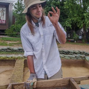 Field school student Aidan Sherwin examines a sherd of ceramic he found while screening through soil excavated south of the Archaearium.