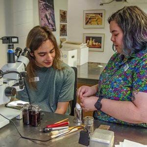 Assistant Conservator Jo Hoppe teaches field school student Aidan Sherwin physical conservation techniques using a microscope.