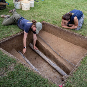 Archaeological Field Technician Ren Willis scores the stratigraphic layers in one of the Smithfield excavation squares as Site Supervisor Anna Shackelford looks on. The pipes traversing the square were installed in the 1930s and the network cables in the 2000s.