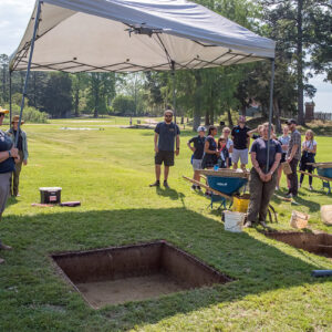 The archaeological crew hold a tent to provide shade for overhead record photography done by drone. The drone is visible at top coming in for the shot.
