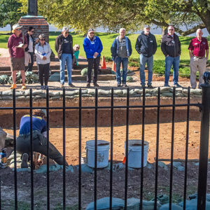 Staff Archaeologist Caitlin Delmas gives an archaeology update to volunteers and staff just outside the Archaearium as Archaeological Field Technician Ren Willis conducts excavations there.