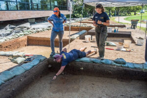 Staff Archaeologist Caitlin Delmas scores a burial with her trowel in preparation for record photography. Senior Staff Archaeologist Mary Anna Richardson and Archaeological Field Technician Eleanor Robb look on.