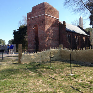 Four grave markers enclosed by partially-reconstructed wall with brick church in background