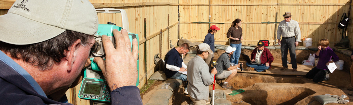 Archaeologist recording with a transit while a group analyzes features in the background