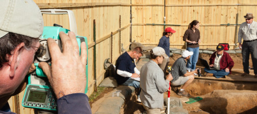 Archaeologist recording with a transit while a group analyzes features in the background