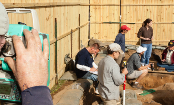 Archaeologist recording with a transit while a group analyzes features in the background