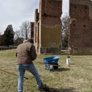 Excavator with wheelbarrow in front of brick ruins