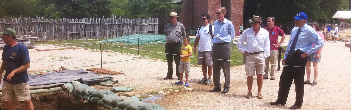 Staff and visitors watch an archaeologist working in an excavation unit