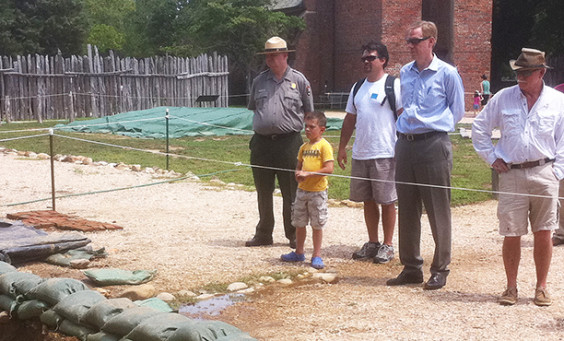 Staff and visitors watch an archaeologist working in an excavation unit