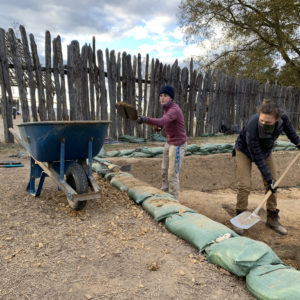 two archaeologists shoveling dirt from a unit into a wheelbarrow