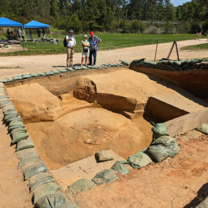 A volunteer describes the well and ditch excavations to some visitors. The excavations to the north are visible in the background.