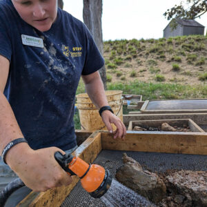 Staff Archaeologist Caitlin Delmas uses water to reveal a sword hilt hidden inside an earthen blob.