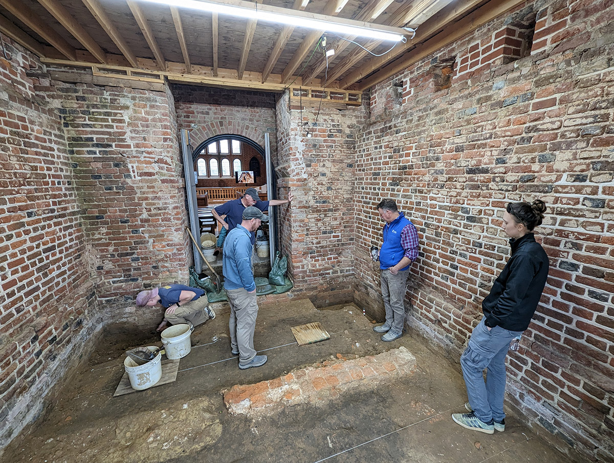 The archaeological team discusses the Church Tower excavations. The foundations of the 1617 church can be seen midway through its footprint. The dark soil is likely the burned remains of the previous tower destroyed during Bacon's Rebellion.