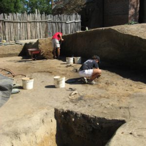 Excavated ditch feature in the foreground with two archaeologists troweling and shoveling in the background