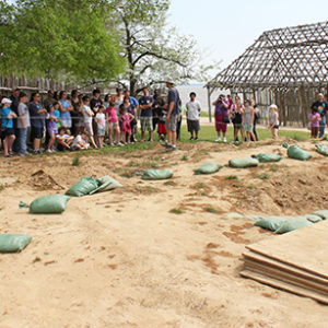 archaeologist giving a tour to a large group of visitors in front of an excavation area and a wooden building frame