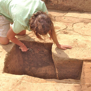 archaeologist bending over an excavation unit and scraping the sides with a trowel