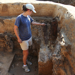 archaeologist points with a trowel to stratigraphic layers in a deep excavation unit