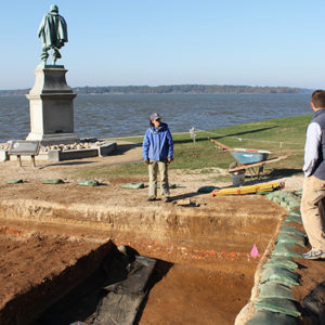 Three archaeologists standing on the edges of an excavation unit lined with sandbags