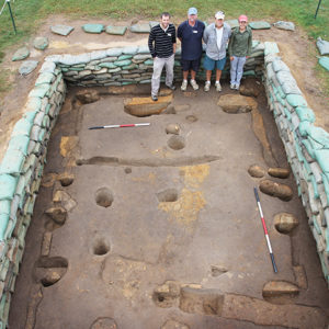 Four archaeologists standing at the edge of an excavation unit filled with a variety of features and lined with sandbags