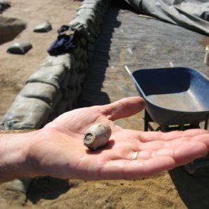 Hand holding pipe bowl with view of wheelbarrow and excavation unit lined with sandbags in the background