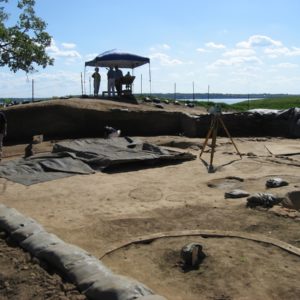 Archaeologists walking around the perimeter of an excavation unit containing a transit and various features