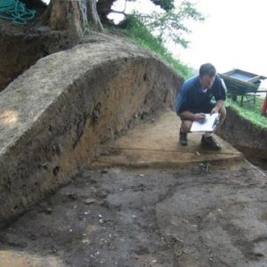 Archaeologist troweling in an excavation unit with a curving wall showing varied stratigraphy