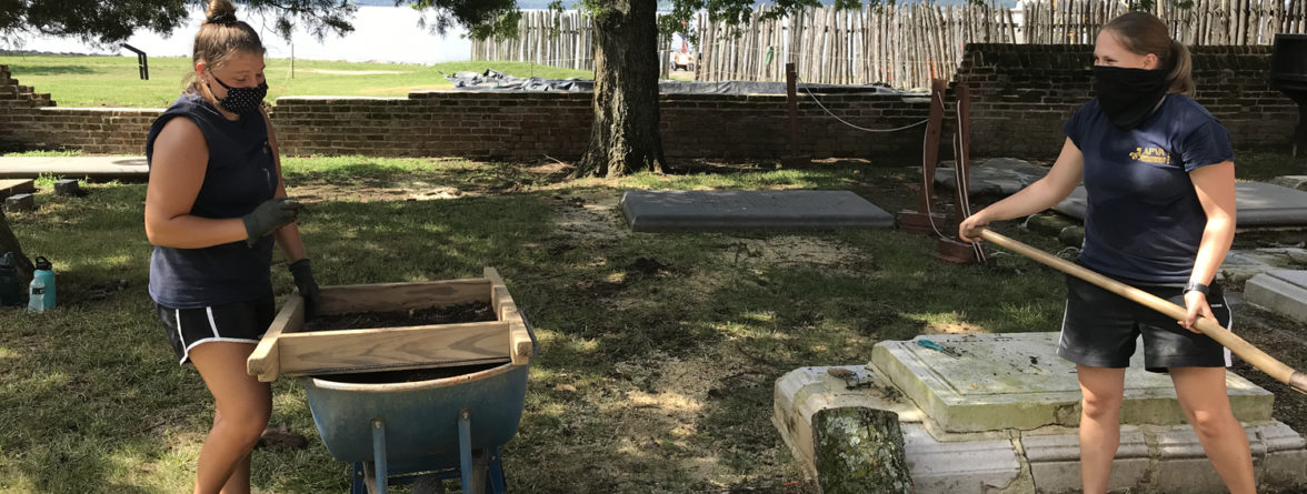 One archaeologist screening artifacts and another excavating with a shovel in a churchyard