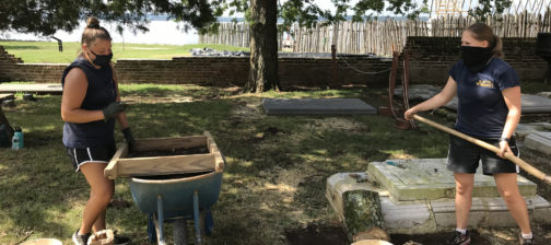 One archaeologist screening artifacts and another excavating with a shovel in a churchyard