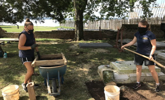 One archaeologist screening artifacts and another excavating with a shovel in a churchyard