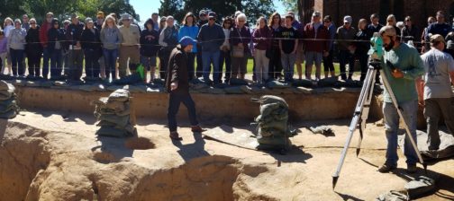 Large group listens to tour guide while archaeologists record finds