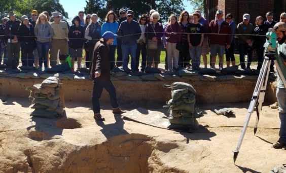 Large group listens to tour guide while archaeologists record finds