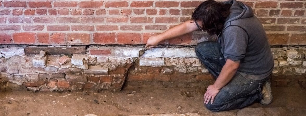 Archaeologist gestures with trowel to brick foundations