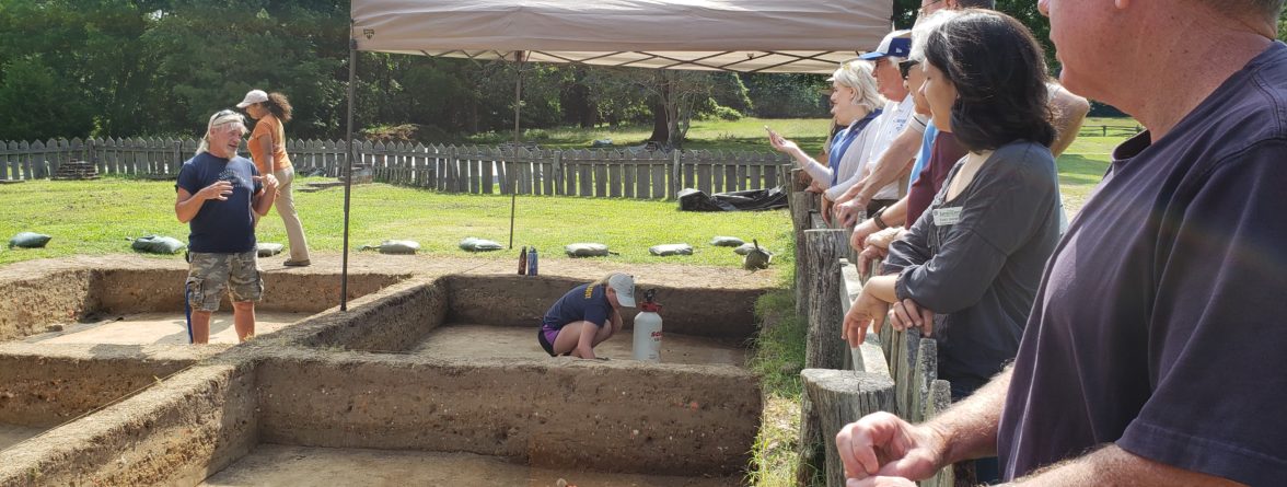 archaeologist standing in an excavation unit and talking to a group of volunteers standing behind a fence