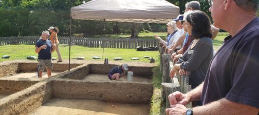 archaeologist standing in an excavation unit and talking to a group of volunteers standing behind a fence