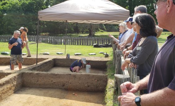 archaeologist standing in an excavation unit and talking to a group of volunteers standing behind a fence