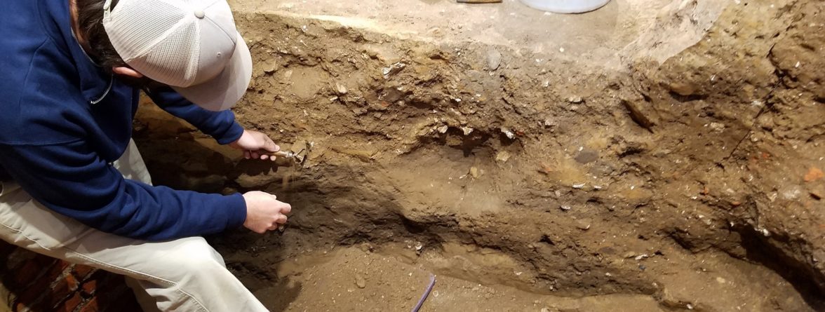 archaeologist using a trowel to scrape soil from a unit wall