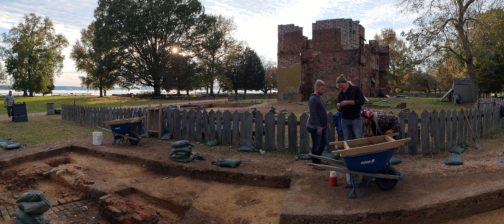 Archaeologists examine artifact while standing in excavations in front of brick ruins