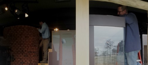 Man standing on a ladder and dusting the top of a museum display next to a window