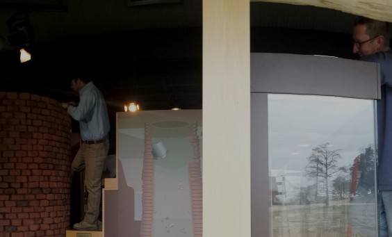 Man standing on a ladder and dusting the top of a museum display next to a window